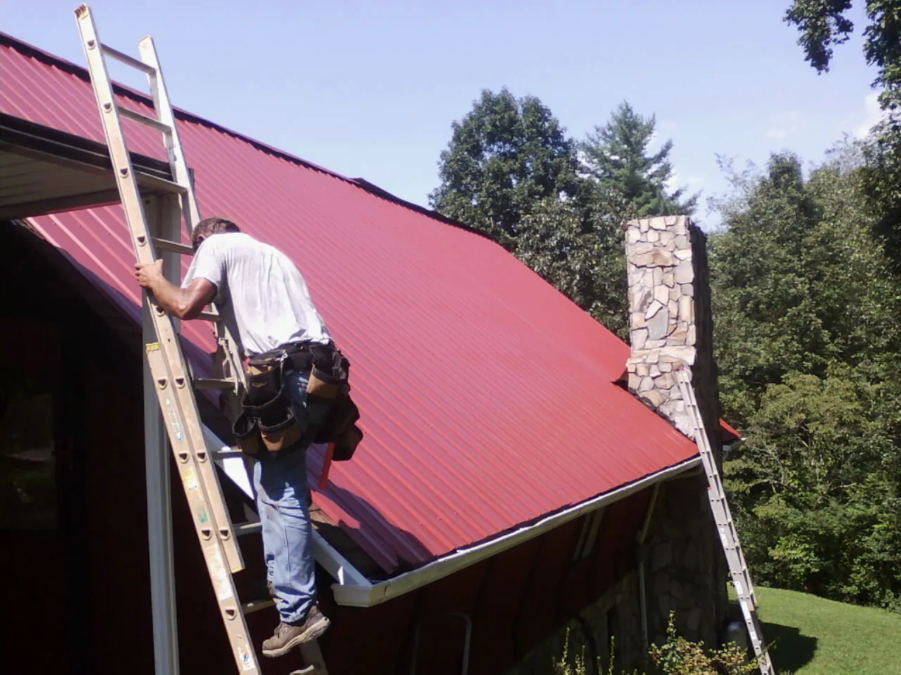Roofer on a ladder beside a red metal roof and stone chimney — Soffit & Fascia Repair by Roofing Brothers in Pavilion